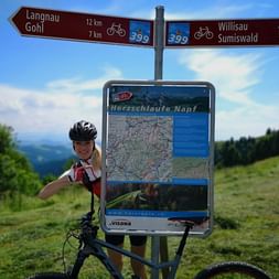 zEine Mountainbikerin steht mit ihrem bike hinter einer Tafel auf der die Karte für die Route Summiswald-Napf abgebildet ist. Oberhalb der Karte sind 2 Wegweiser, die rechts den Wanderweg nach Willisau/Summiswald anzeigen und links nach Langnau/Gohl.