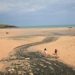 Die Harlyn-Bay. Ein riesen Sandstrand an dem Kinder spielen und verschiedene Menschen spazieren. Hinten links im Bild sieht ma noch einen kleinen Grashügel mit einem Häuschen und am Horizont ein bedekter grauer Himmel.