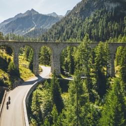 Two cyclists on a winding mountain road beneath a stone railway viaduct in Graubünden. Dense forest covers the mountainsides.