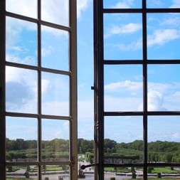 Blick durch hohe Palastfenster in Drottningholm auf formale Barockgärten mit geometrischen Rasenflächen, geschnittenen Hecken und einem See.