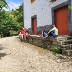 Vier Wanderer rasten auf Steinstufen eines weißen Gebäudes mit roten Türen bei Rundwanderung nahe Pinhão. Kopfsteinpflaster und Weinberge sichtbar.