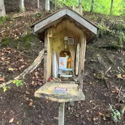 Kleiner Holzschrein auf einem Pfosten im Wald mit einer Schnapsflasche, einem Thermometer und einem Gästebuch auf einem Holzbrett.