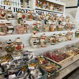Ausstellung handbemalter portugiesischer Keramik-Honigtöpfe und Krüge auf dem Markt von Olhão. Regale zeigen bunte Töpferwaren mit traditionellen Motiven über Trockenfrüchten.