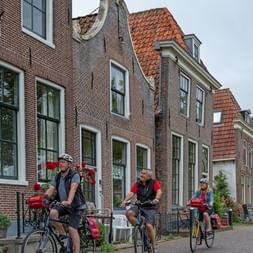 Trois cyclistes avec vélos de randonnée font une pause sur une rue pavée bordée de maisons hollandaises traditionnelles en brique.