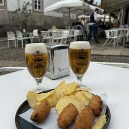 Zwei Estrella Galicia Biere mit frittierten Snacks und Chips auf einem Teller auf einer Außenterrasse in Minho-Porto. Weiße Tische und Sonnenschirme sichtbar.