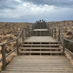 Holzsteg mit Treppe über Sanddünen bei Lagoa dos Salgados. Drei Personen sind oben unter bewölktem Himmel sichtbar.