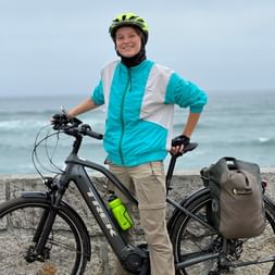 Lächelnde Radfahrerin Nathalie mit gelbem Helm und türkisfarbener Jacke steht mit schwarzem E-Bike an Küstenpromenade mit Blick auf den Atlantik.