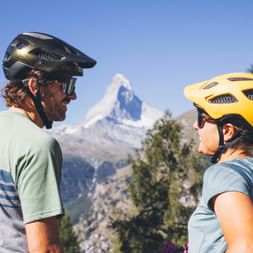Two cyclists stand in front of the Matterhorn and look at each other.