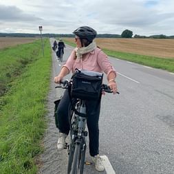 Radfahrerin in rosa Jacke mit Helm auf asphaltierter Straße durch Agrarlandschaft. Zwei weitere Radfahrer voraus sichtbar.
