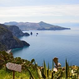 Panoramablick auf die Liparischen Inseln mit dramatischen Klippen, türkisfarbenem Mittelmeer und Felstürmen. Vegetation und ein Schild sind im Vordergrund sichtbar.