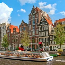 Bateau touristique sur un canal d'Amsterdam passant devant des maisons hollandaises à pignons colorées sous un ciel bleu.