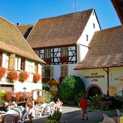 Charmante place de village alsacien avec maisons à colombages, jardinières rouges, terrasse de café et visiteurs profitant du soleil.
