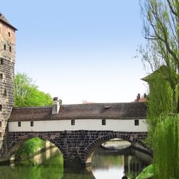 Historischer Henkersteg in Nürnberg mit Steinbögen über dem Fluss, mittelalterlichem Turm, Fachwerkhäusern und Trauerweide.