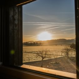 Blick durch ein Planwagenfenster in Courtedoux, Jura Ajoie, mit Sonnenaufgang über frostbedeckten Feldern mit kahlen Bäumen und fernen Hügeln.