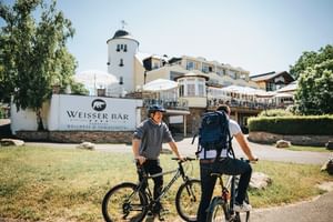 Zwei Radfahrer mit Fahrrädern vor dem Hotel Weisser Bär, einem weißen Gebäudekomplex mit markanten runden Turm und Außenterrasse.