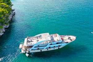 Luftaufnahme der MS Olmissum, ein weißes Kreuzfahrtschiff mit blauem Streifen, vor Anker in türkisem Wasser nahe bewaldeter Küste.