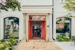 Eleganter Hoteleingang mit orangenen Vorhängen, Steinsäulen und gewölbten Fenstern. Zickzack-Steinpflaster führt zur offenen Tür.