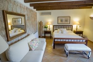 Chambre double spacieuse avec mur en pierre, poutres en bois, canapé blanc et lit en bois foncé. Sol en carreaux de terre cuite.