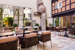 Außenterrasse im Hotel Le Colombier mit orangefarbenen Polsterstühlen, runden Tischen und dekorativer Steinskulptur umgeben von historischen Gebäuden.