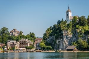 Panoramablick auf Traunkirchen mit traditionellen Gebäuden am Seeufer und einer weißen Kirche mit Uhrturm auf einem Felsen.
