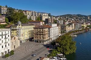 Luftaufnahme der Uferpromenade von Lugano mit Hotel International zwischen bunten Gebäuden am See, Bergen im Hintergrund, klarer blauer Himmel.
