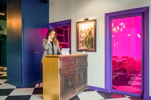 Moderne Hotelrezeption mit Schachbrettboden, Holztheke und Rezeptionistin am Telefon. Lila gerahmte Tür zeigt rosa beleuchteten Raum.