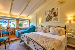 Chambre double lumineuse avec lit en fer forgé, poutres apparentes, murs en pierre, fauteuil turquoise et œuvre d'art au-dessus du lit à Son Sant Jordi Pollenca.