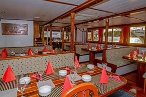 Innenraum des MS Macek Schiffsrestaurants mit Holztischen, roten Servietten, Weingläsern und gestreiften Tischsets. Sitzbänke und Fenster säumen die Wände.