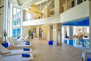 Moderner Hallenbad-Bereich mit weißen Liegen, blauen Handtüchern und großen Fenstern. Zweistöckiger Raum mit Empore im Salzburger Hof Bad Gastein.