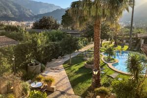 Vue du jardin à Can Quatre à Sóller avec piscine incurvée, palmiers, pelouse et montagnes de Tramuntana en arrière-plan sous ciel bleu.