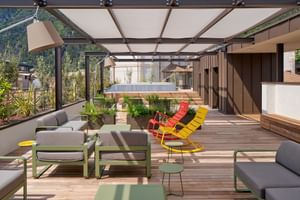 Geräumige Dachterrasse mit Holzdielen, grauen Sofas, bunten Stühlen in Rot und Gelb, Glasüberdachung und Bergblick im Hintergrund.