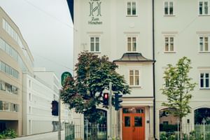 Weißes historisches Hotel zum Hirschen Gebäude in Salzburg mit verziertem Eingang, grünen Bäumen und modernen weißen Gebäuden links.