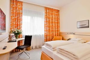 Modernes Hotel-Doppelzimmer mit orangen Vorhängen, zwei Einzelbetten, Holzschreibtisch mit schwarzem Stuhl und gerahmten Bildern.