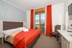 Modernes Hotelzimmer mit Doppelbett, oranger Tagesdecke und Vorhängen, Balkontür mit Bergblick, braunem Kopfteil und Schreibtisch in Malcesine.
