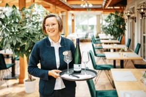 Lächelnde Mitarbeiterin in dunklem Blazer hält Tablett mit Weinflasche und Gläsern in hellem Restaurant mit Holzbalken und grünen Stühlen.
