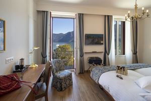 Helles Doppelzimmer mit Seeblick im Hotel International Lugano. Holzboden, gemusterter Sessel, Schreibtisch und Balkontür mit Blick auf Berge und Wasser.