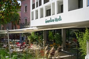 Modernes weißes Hotelgebäude mit Giardino Verdi Schild, mit überdachter Terrasse mit bunten Stühlen und üppiger Gartenlandschaft.