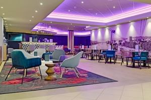 Moderne Hotelbar mit violetter Deckenbeleuchtung, bunten Sesseln, Esstischen und Wandgemälde. Zeitgenössische Möbel und Ambientebeleuchtung.