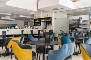 Modernes Restaurant-Interieur im Hotel Jupiter Lisboa mit bunten Stühlen in Gelb und Blau um dunkle Tische, weiße Bar mit Flaschen.