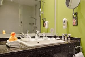 Modernes Hotelbadezimmer mit hellgrüner Akzentwand, weißem Waschbecken auf dunkler Granitplatte, Glasdusche, Vergrößerungsspiegel und Quietscheente.