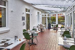 Überdachte Terrasse im Hotel Upstalsboom mit Ziegelboden, grünen Stühlen, weißen Tischen und Glaswänden mit Blick auf den Außenbereich.