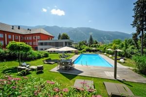 Außenpool im Hotel Ertl in Spittal mit Liegestühlen, Sonnenschirmen und angelegten Gärten. Rotes Hotelgebäude und Berge im Hintergrund sichtbar.