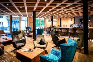 Salon d'hôtel moderne avec plafond à poutres apparentes, fauteuils colorés, tables en bois et bar avec tabourets en arrière-plan.