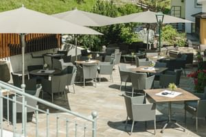 Terrasse extérieure du Schwarzer Adler St. Anton avec chaises grises, tables en bois sous grands parasols, sol en pierre et paysage montagnard.