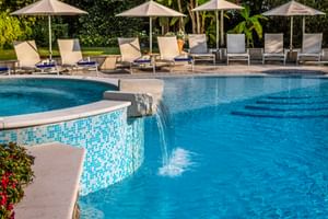 Blauer Swimmingpool im Regina Adelaide Garda Hotel mit Wasserspiel, beigen Liegestühlen unter Sonnenschirmen und üppiger Vegetation.