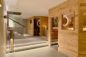 Modern spa area with wooden sauna cabins featuring circular windows, illuminated steps, and light wood paneling in a hotel wellness facility.