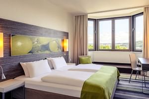 Modernes Doppelzimmer im Mercure Koblenz mit zwei Betten, grünen Akzenten, Holzkopfteil, großen Fenstern mit Stadtblick und zeitgemäßer Einrichtung.