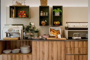 Frühstücksbuffet mit Brotkörben, frischem Obst und Gebäck auf Holztheke. Wandregale zeigen Dekoartikel und Pflanzen.