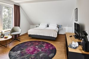 Modernes Hotelzimmer mit Doppelbett, Holzboden, Dachschräge, rundem lila Teppich, Schreibtisch mit Kaffeemaschine und Fensterblick.