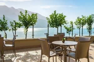 Außenterrasse der Villa Dirce Limone mit beigen Stühlen und runden Tischen, kleinen Topfbäumen, mit Blick auf einen See und Bergen im Hintergrund.